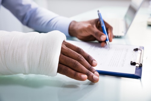 Injured Man With Bandage Hand Filling Insurance Claim Form On Clipboard